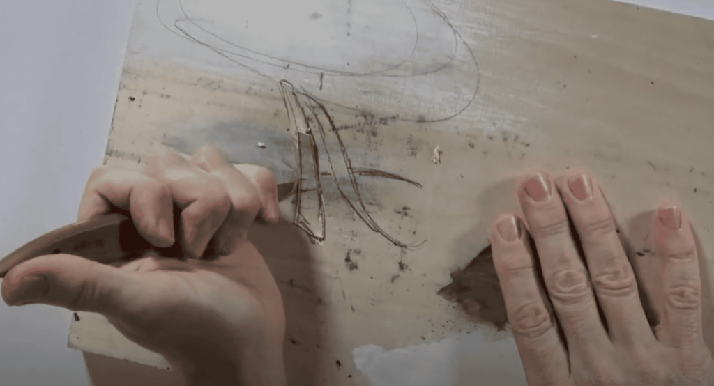 A person is using a carving knife to create a design in wood on a workbench, showing the hands gripping the tool and the outlines on the wood. This image captures the art of wood carving, a skill taught in wood carving classes and lessons.